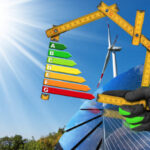 Hand with work glove holding a wooden folding ruler in the shape of house with energy efficiency rating on a blue sky with a solar panel and wind turbine