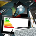 Model house with a light bulb and a energy efficiency rating on a desk with a tablet pc, computer keyboard and work tools. Project of ecological house concept